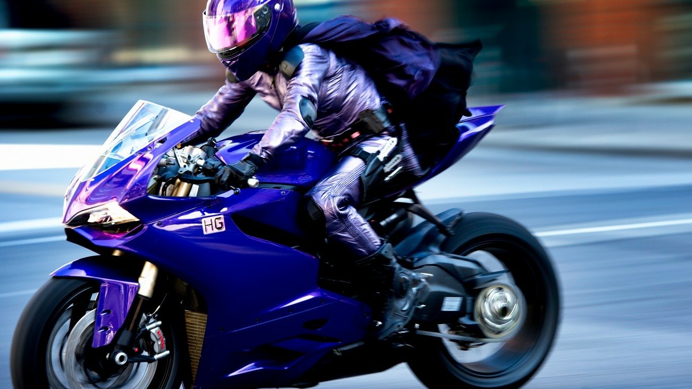 Costumed motorcyclist from the movie Kick-Ass 2 speeds on a purple sportbike, cape and helmet blurred by motion.