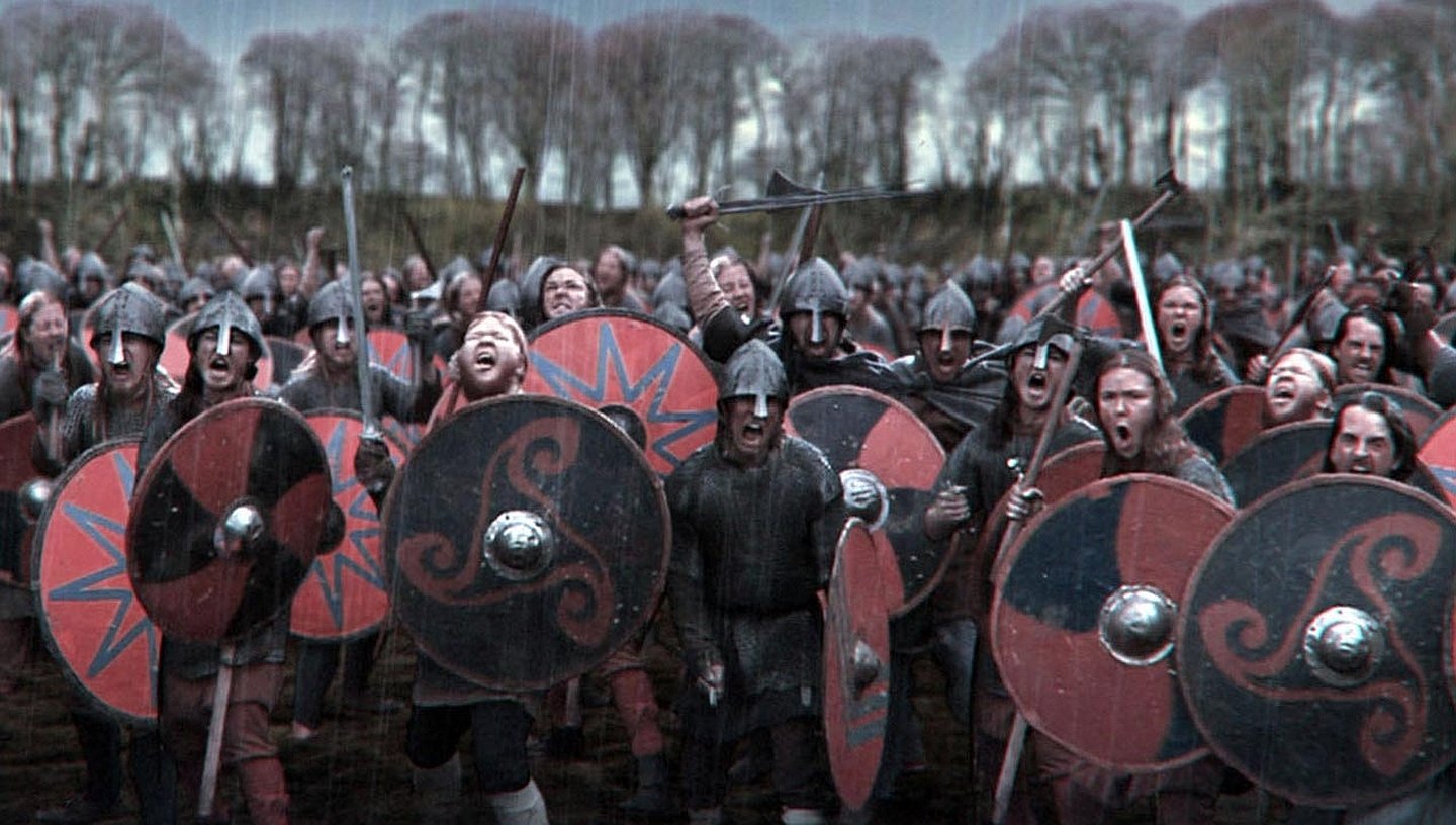 A large group of Vikings warriors from the TV show Vikings (2013) stand ready in formation, holding round shields with various red, black, and silver designs.