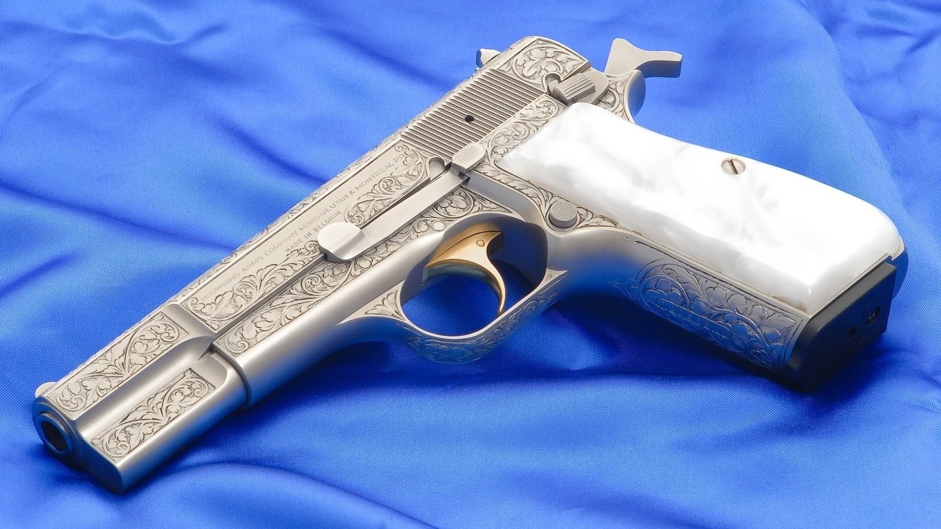 A detailed Browning pistol with an intricately engraved silver finish and a white grip rests on a blue fabric background.