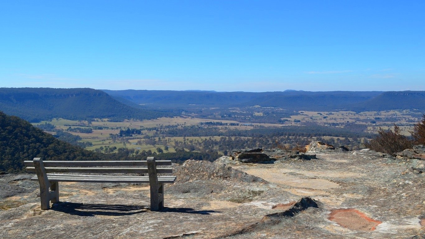 A wooden bench at Hassans Walls Lookout in the Blue Mountains, Australia, overlooking a vast mountain landscape under a clear blue sky.