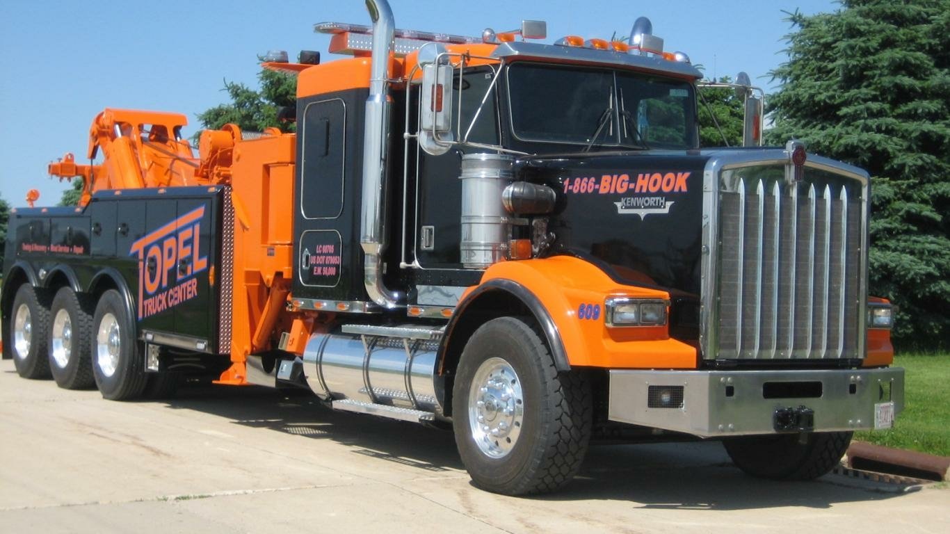 A Kenworth tow truck in bright orange and black, parked on a paved surface, showcasing its robust build and towing equipment against a backdrop of greenery.