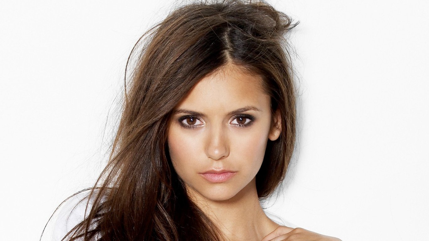 A close-up portrait of celebrity Nina Dobrev, showcasing her long, flowing hair and striking facial features against a plain white background.