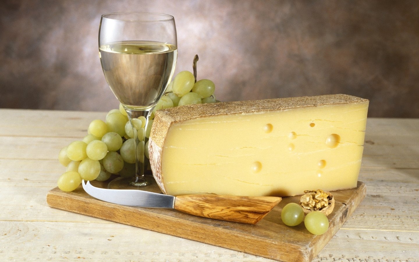 A wedge of Swiss cheese on a wooden board with green grapes, a knife, walnuts, and a glass of white wine.