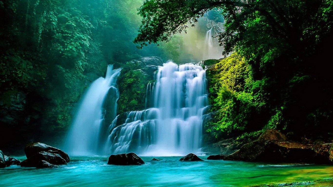A serene waterfall cascades over lush rocks into turquoise waters, surrounded by dense green foliage, creating a tranquil nature scene.