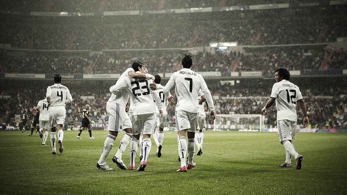 Real Madrid C.F. players celebrate a goal on the pitch, surrounded by a cheering crowd in a high-energy sports atmosphere.