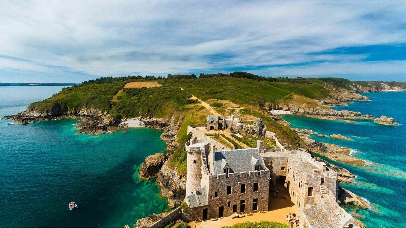 Aerial view of the man-made Fort-la-Latte perched on a rocky coastline surrounded by blue ocean waters and green landscape under a partly cloudy sky.