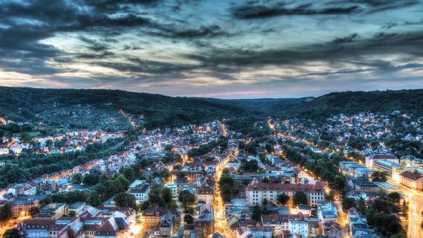 Aerial view of Jena, Germany at dusk: illuminated streets winding through a valley of tightly packed houses and wooded hills.