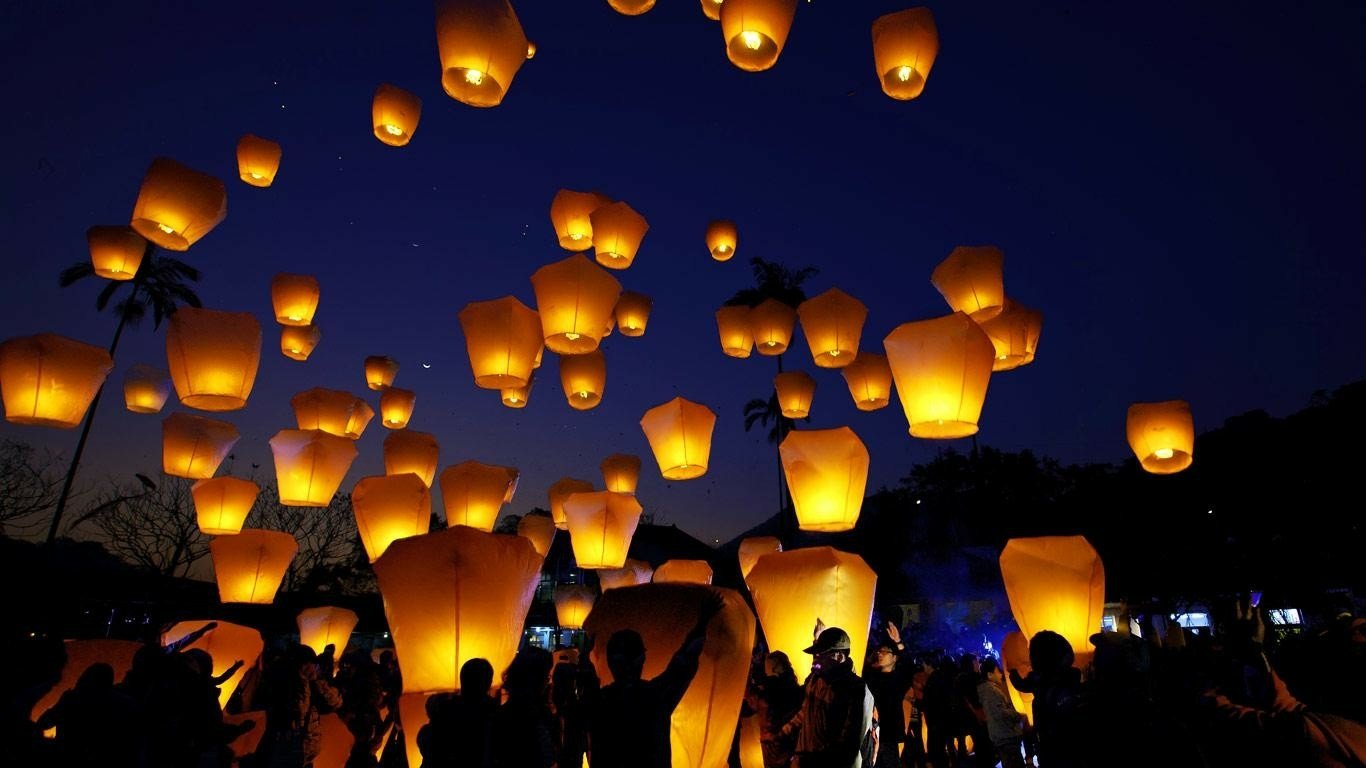 A crowd releases numerous glowing man-made lanterns into the night sky, creating a warm, illuminated display against the dark blue backdrop.