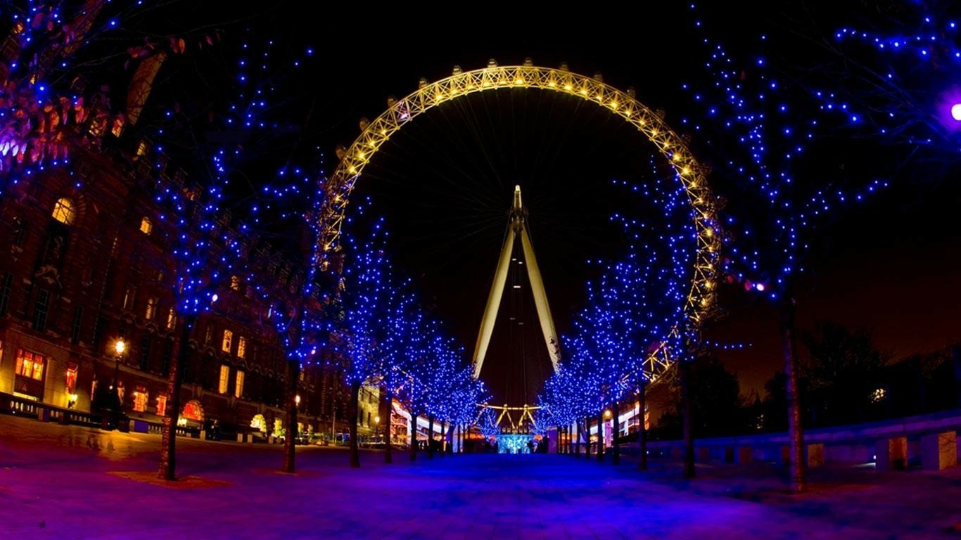 The illuminated London Eye stands tall at night, framed by trees adorned with blue lights along the riverside walkway.