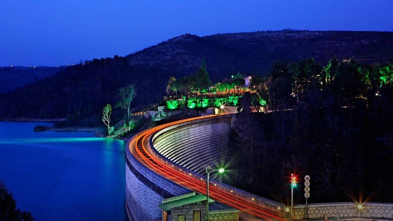 Man-made Marathon Dam at night, its curved concrete wall lit with green lights, water on one side and streaking vehicle light trails along the roadway.