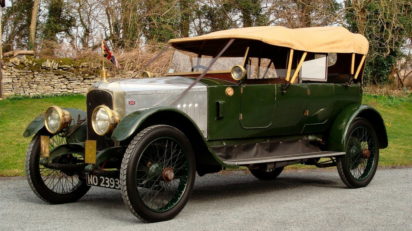 A vintage green and silver Vauxhall vehicle with a beige soft top parked on a paved road beside a stone wall and trees.