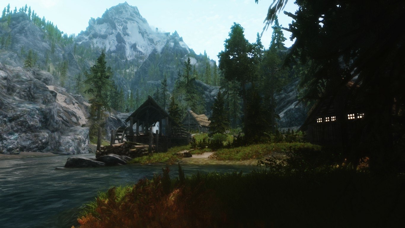 A serene lakeside village set against towering pine trees and rugged mountains in the natural world of The Elder Scrolls V: Skyrim video game.