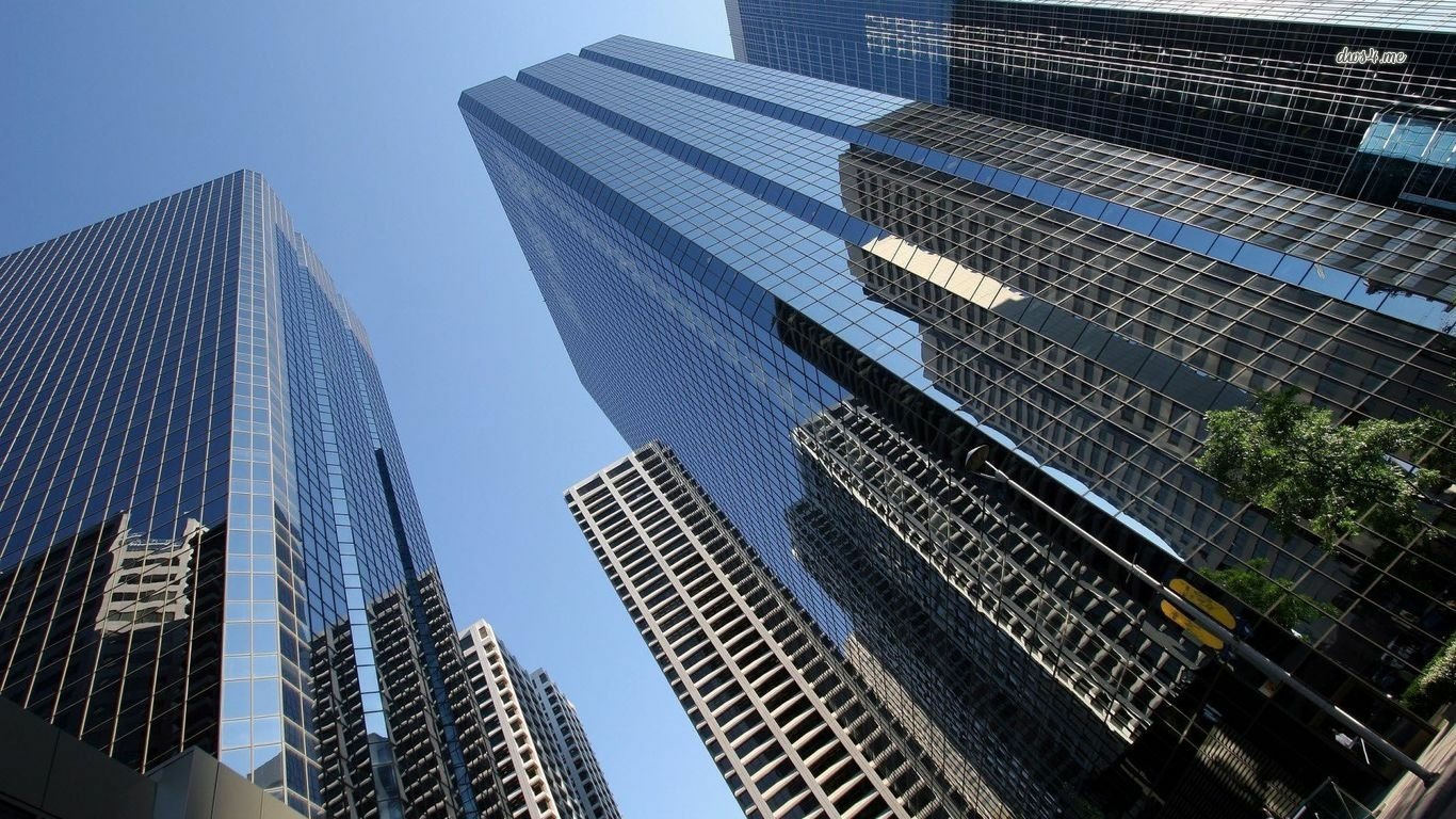 A view looking up at tall, man-made glass skyscrapers in New York against a clear blue sky.