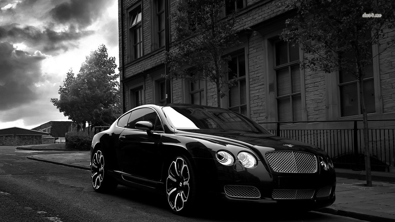 A sleek black Bentley Continental GT parked on a quiet street with historic buildings and dramatic clouds in the background.