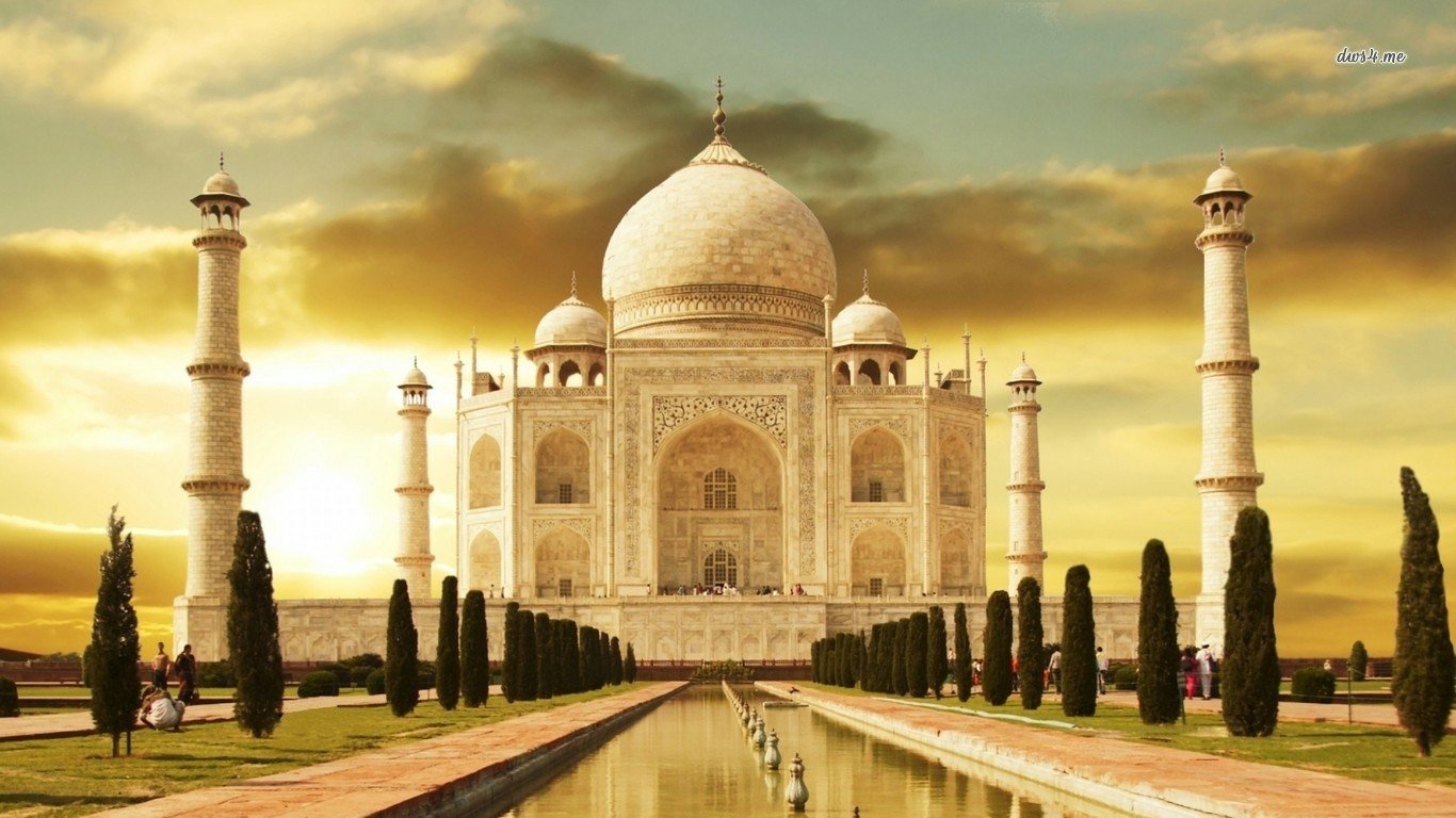 A stunning view of the Taj Mahal, a renowned man-made masterpiece, features its iconic dome and minarets against a golden sunset, surrounded by lush greenery and a reflective waterbody.