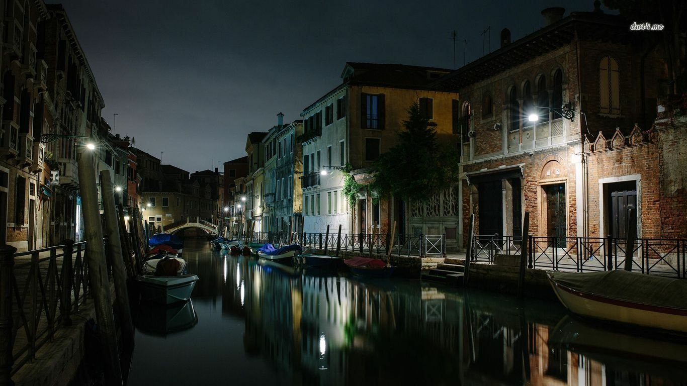 Night view of a man-made canal in Venice with moored boats, illuminated historic buildings and glassy reflections.