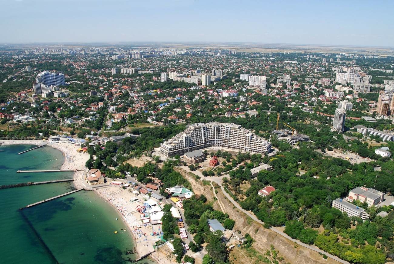 Aerial view of a man-made coastal area in Odessa, featuring beachfront structures, green spaces, and city buildings extending into the horizon.