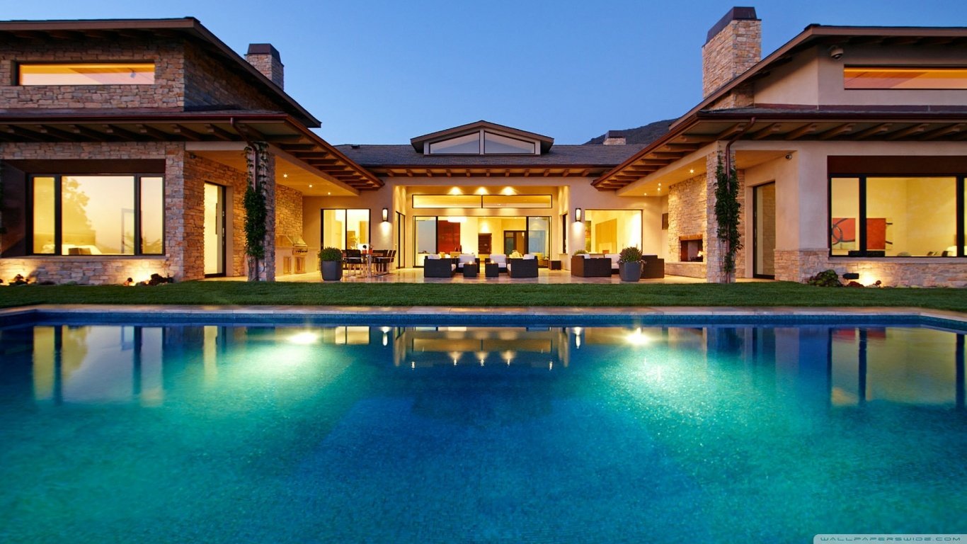 A modern house with large windows and stone details overlooks a sparkling pool, illuminated by lights at dusk, creating a serene and inviting atmosphere.