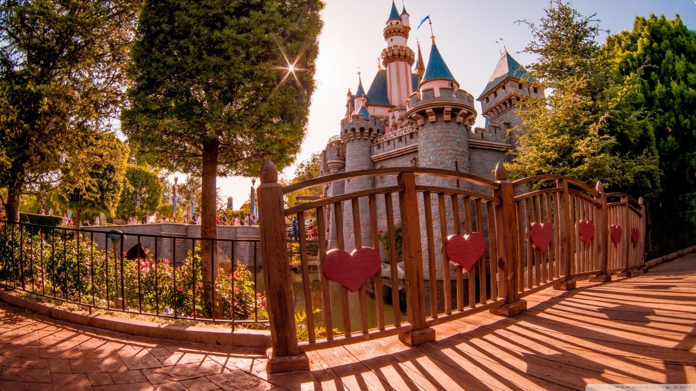 Sunset view of Disneyland's iconic Disney castle behind a wooden fence decorated with red hearts, surrounded by trees and warm golden light.