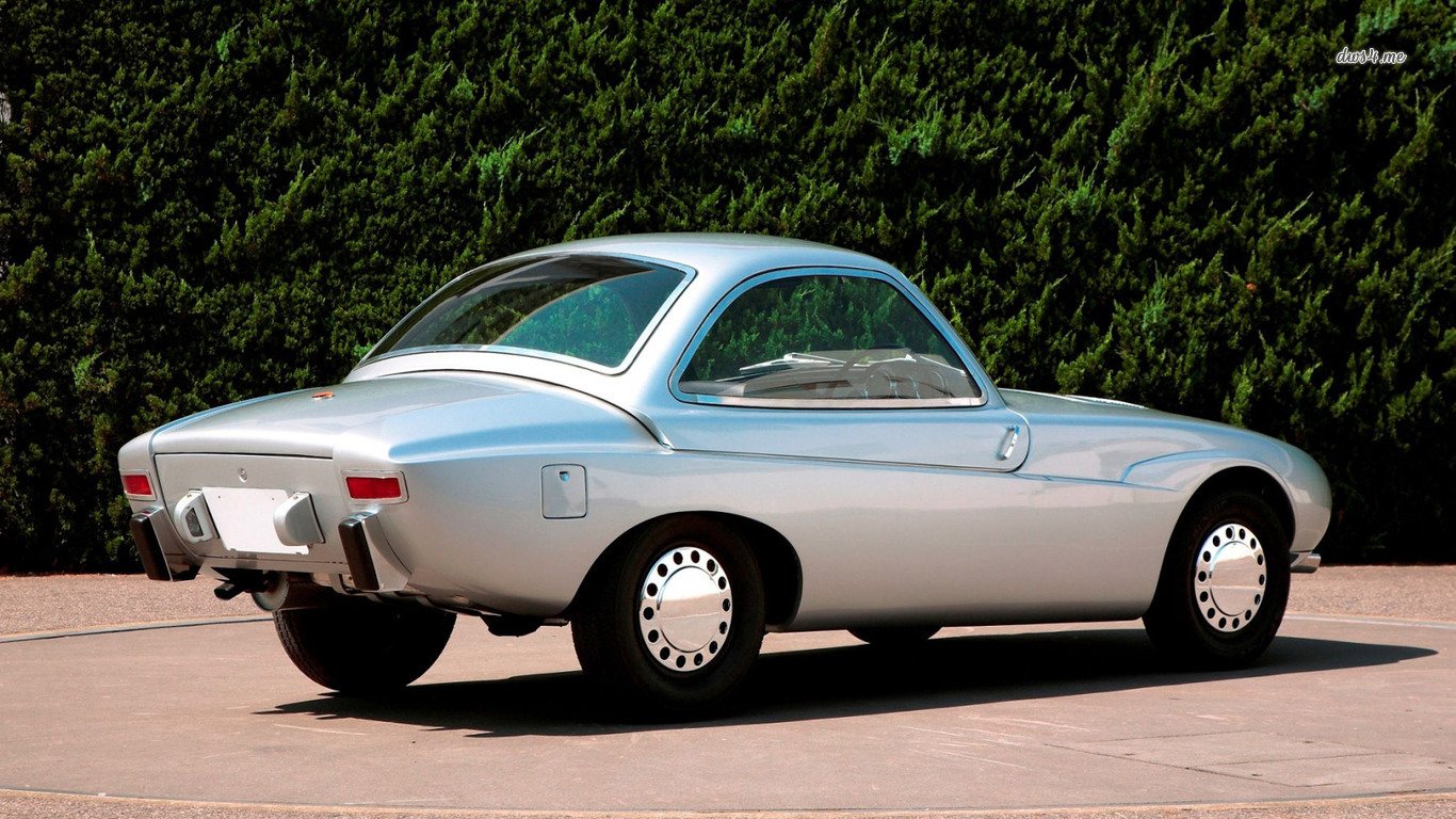 Silver Toyota Publica vehicle — compact vintage coupe shown in a rear three-quarter view, parked on a driveway with tall green hedges in the background.