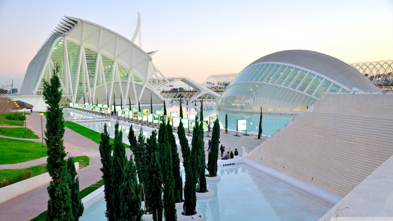Man-made futuristic white architecture: curved glass-and-steel buildings around reflecting pools, framed by cypress trees under a pastel sunset.