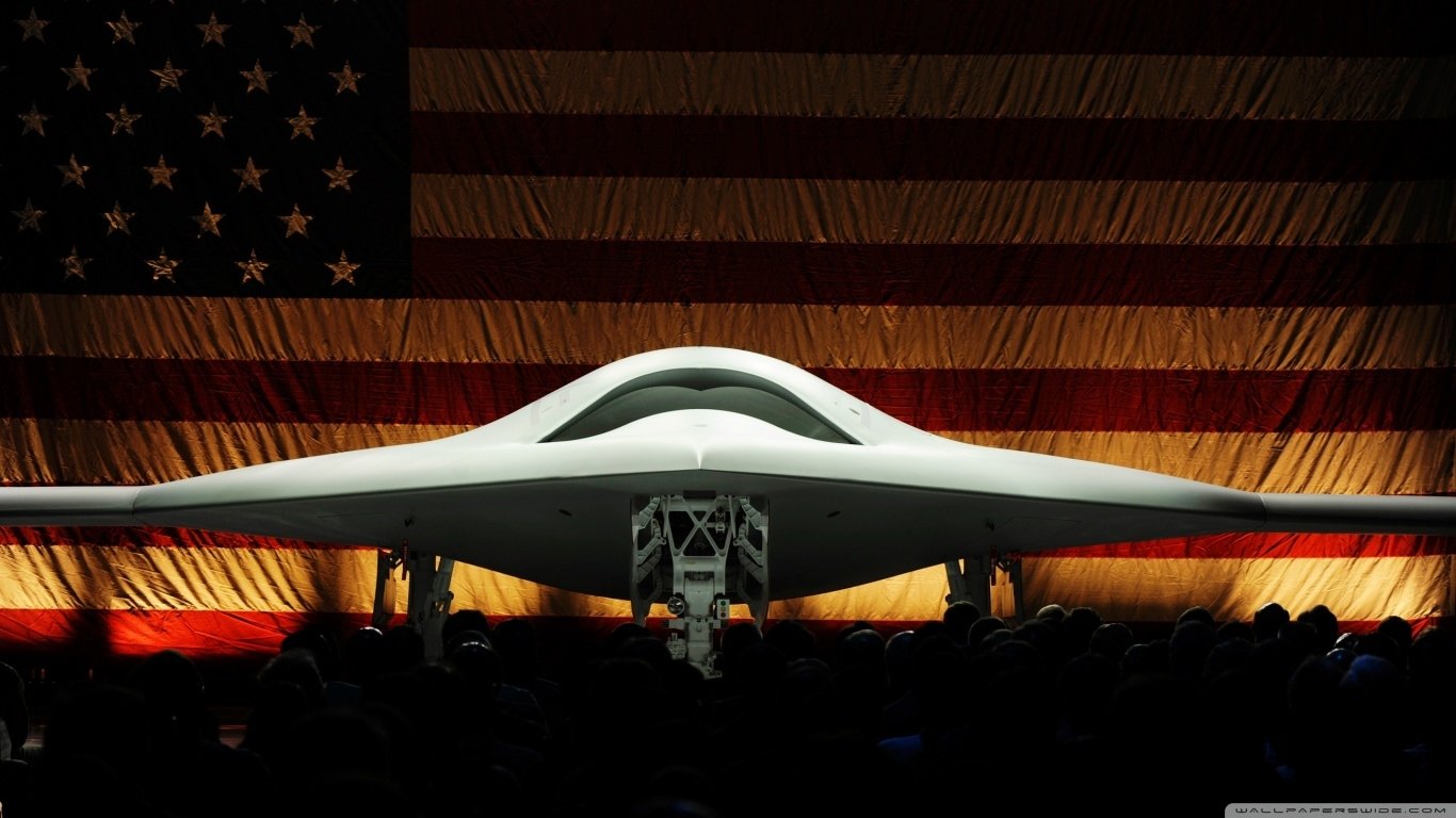 Front low-angle view of a Northrop Grumman X-47B military unmanned aircraft showcased against a large American flag backdrop.