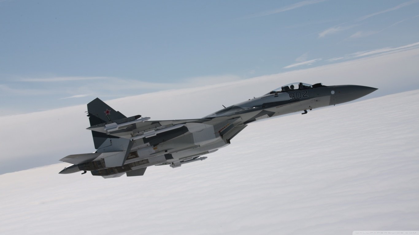A military Sukhoi Su-35 fighter jet soaring above the clouds under a clear blue sky.