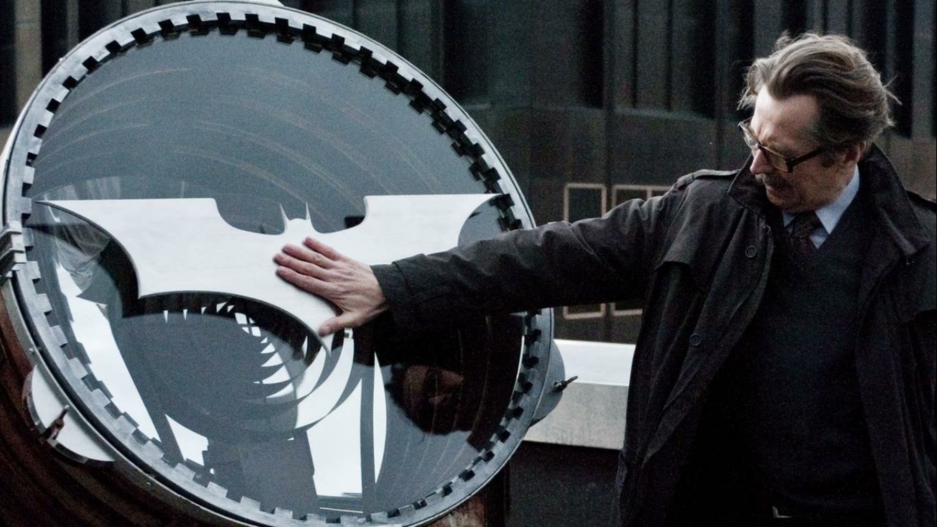 Bespectacled man in a coat reaches to touch the illuminated Bat-Signal on a rooftop — scene from The Dark Knight Rises.