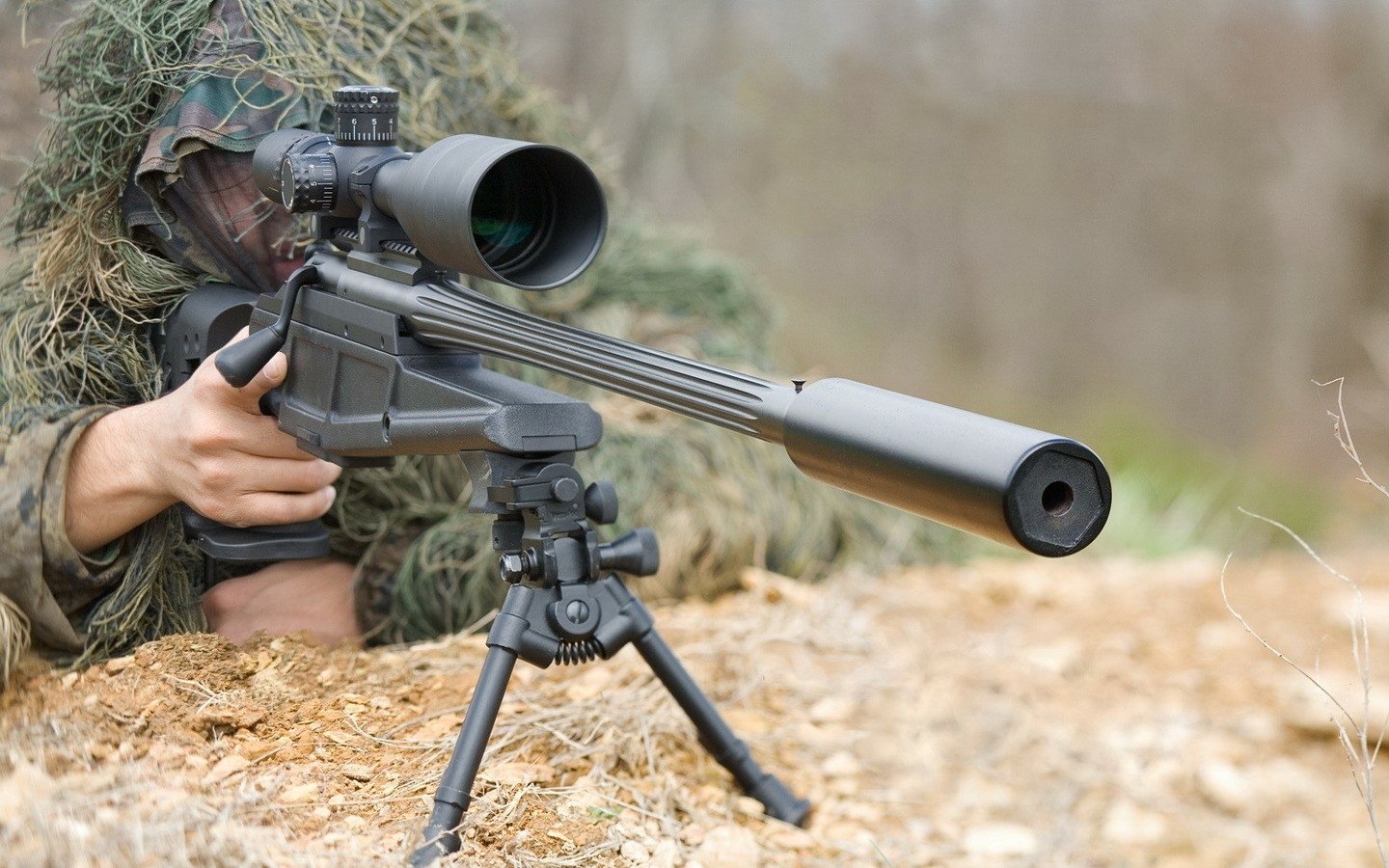 A soldier in camouflage maneuvers while aiming a man-made sniper rifle, equipped with a scope and mounted on a tripod, amidst a natural setting.