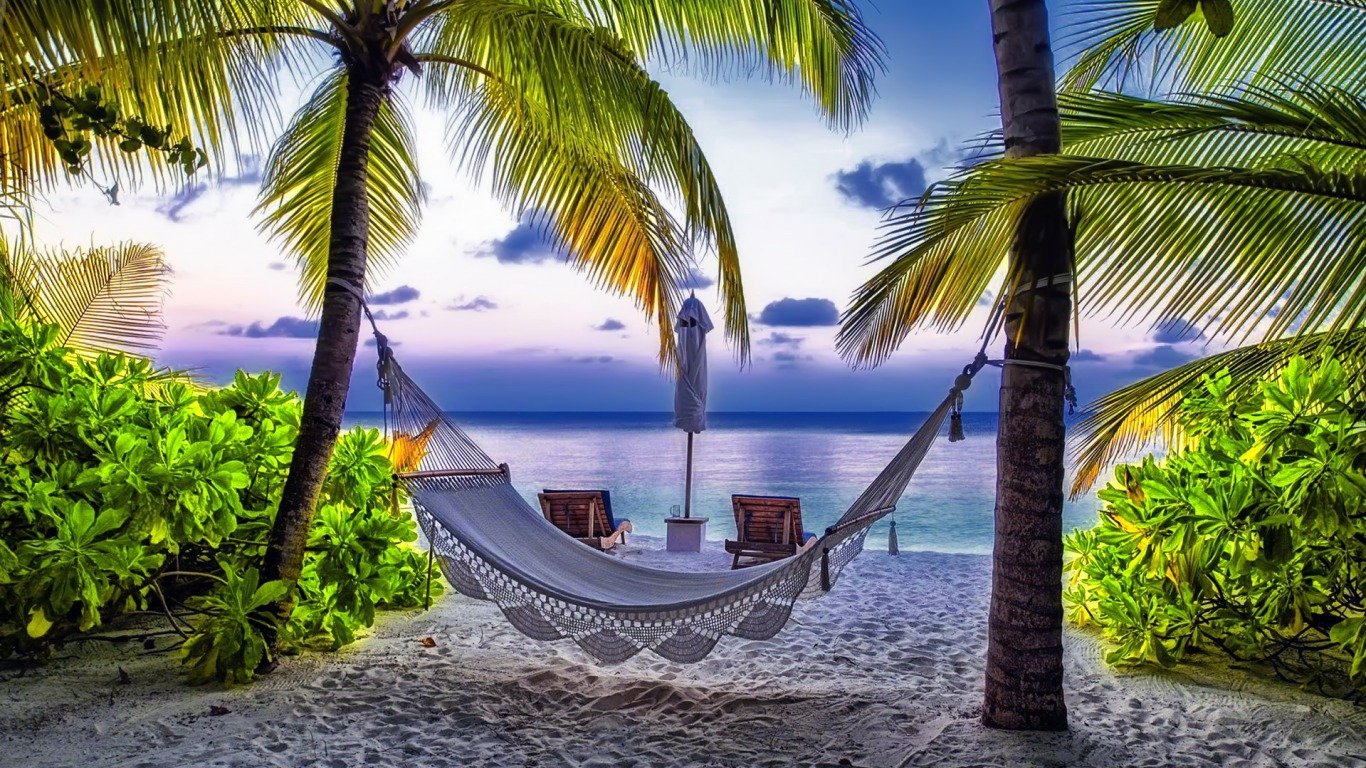 A serene beach scene at sunset, featuring a hammock swaying between palm trees with chairs nearby, framed by lush greenery and tranquil sea views.