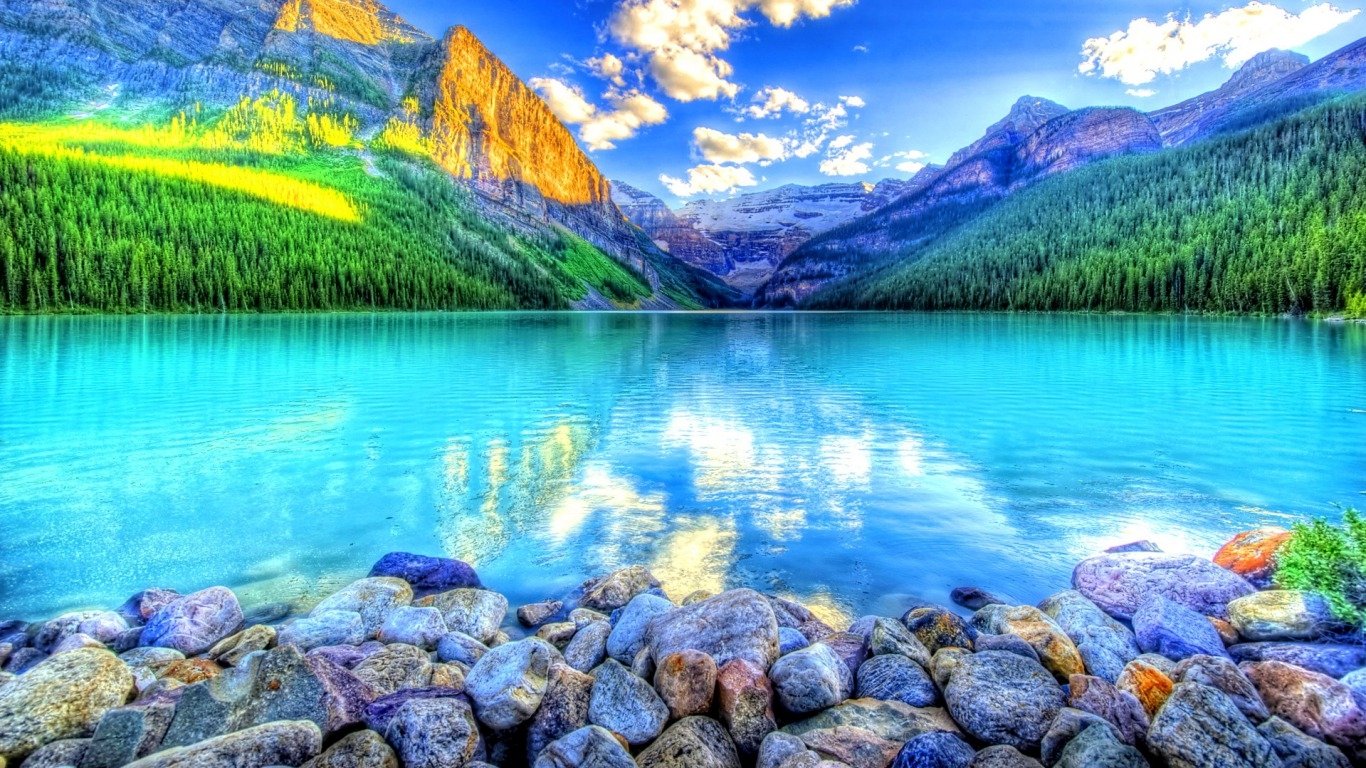 A stunning HDR photograph of a serene lake surrounded by vibrant green forests and majestic mountains, reflecting a bright blue sky with fluffy clouds. Rocks line the shore.