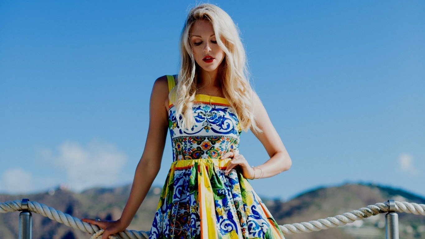 Woman Shea Marie stands outdoors against a clear blue sky, wearing a colorful patterned dress with a belted waist, and looking down thoughtfully.