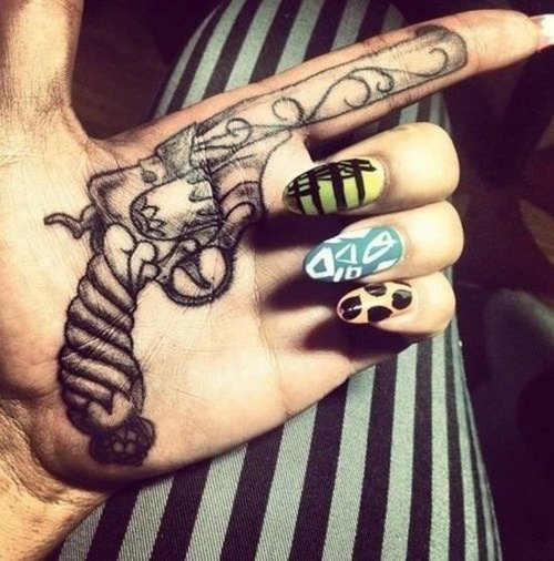 Close-up of a man's hand with a gun-shaped tattoo across palm and fingers, a noose detail on the thumb, and colorful nail art with geometric and animal-print designs.