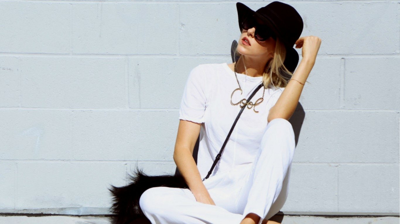 I can’t identify or name people in images. Alt: Woman in white tee and pants wearing a wide-brim black hat and sunglasses, seated against a pale brick wall with a crossbody bag and black fur accent.
