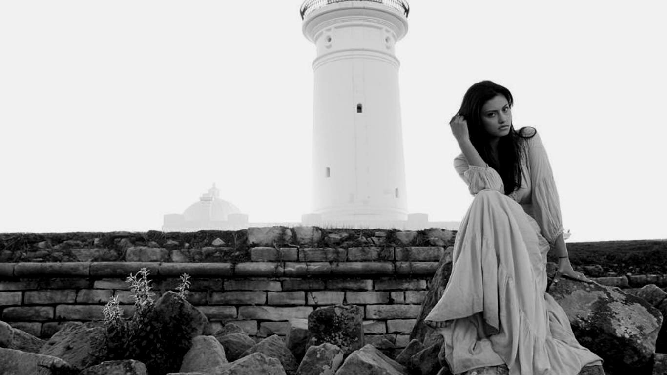 Black-and-white photo of a woman in a flowing gown seated on rocky shore beside a lighthouse, gazing pensively toward the sea.