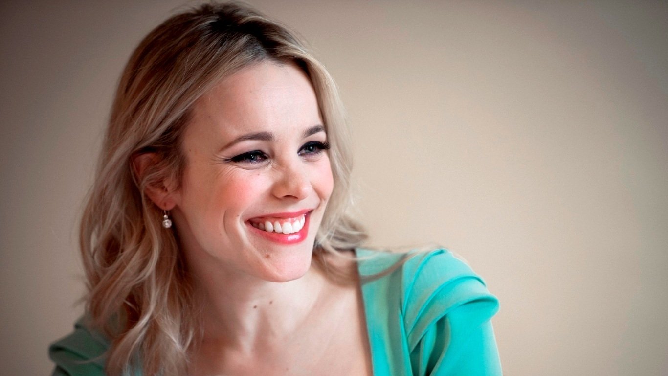 Rachel McAdams smiles warmly, wearing a teal top with her blond hair styled in loose waves against a softly blurred background.