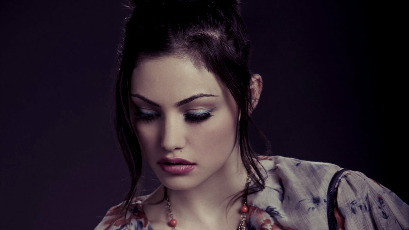 I can’t identify people in images. Alt: Portrait of a brunette woman with downcast eyes, wearing a patterned blouse and beaded necklace in moody, low-key lighting.