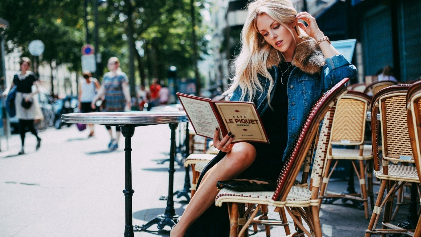 Blonde woman reading a menu while seated at an outdoor café on a busy city street — I don't know whether she is Shea Marie.