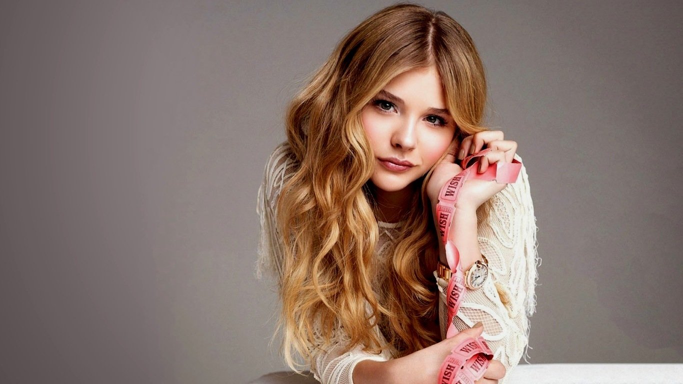Portrait of celebrity Chloë Grace Moretz with long wavy hair, wearing a fringed white top, posing against a gray background.
