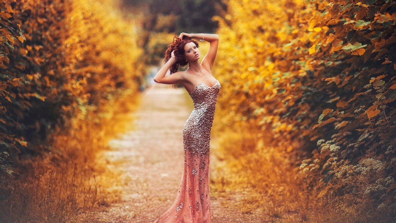 A woman in a shimmering gown poses gracefully on a path surrounded by vibrant autumn foliage in this striking photography.