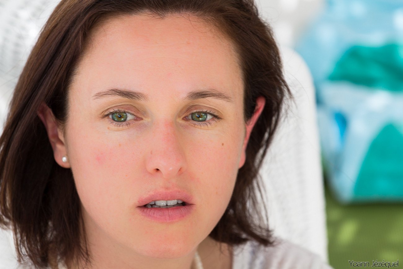 Close-up of a woman’s face with light skin, green eyes, and brown hair, showing a neutral expression against a softly blurred background.