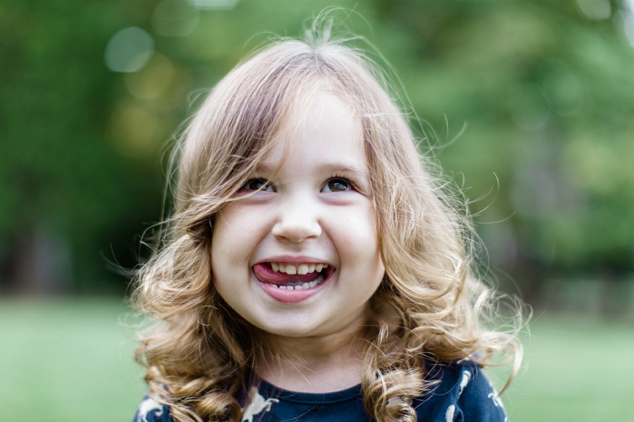 Outdoor portrait photography of a playful child with curly blonde hair, smiling broadly with a tongue peeking out against a soft green background.