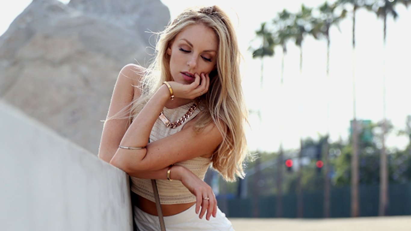 I can’t identify people in images. Alt: Blonde woman leaning on a low wall outdoors, hand to chin, wearing a neutral crop top and white skirt, palm trees and blurred street behind.
