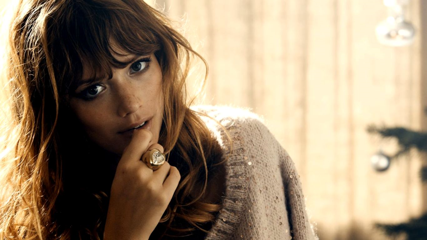 Close-up of an unknown woman with tousled hair, finger to her lips, wearing a knit sweater and statement ring; image tagged Esther Simonet.