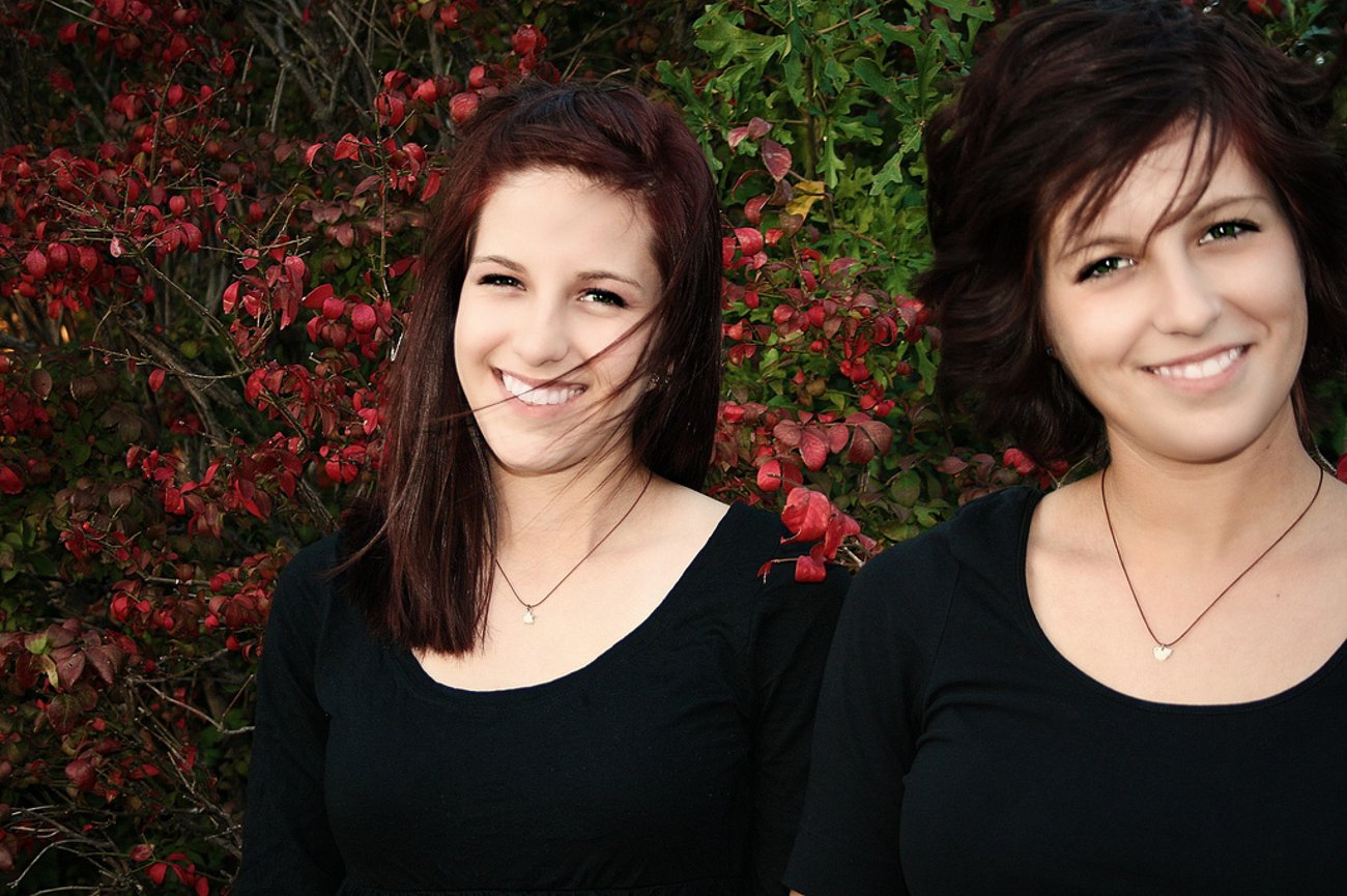Two women standing close in front of red foliage, smiling warmly and sharing a loving, joyful moment.