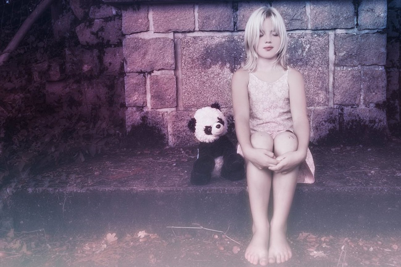 A photography image of a young blonde child sitting barefoot in front of a stone wall, with a panda stuffed animal placed beside her.