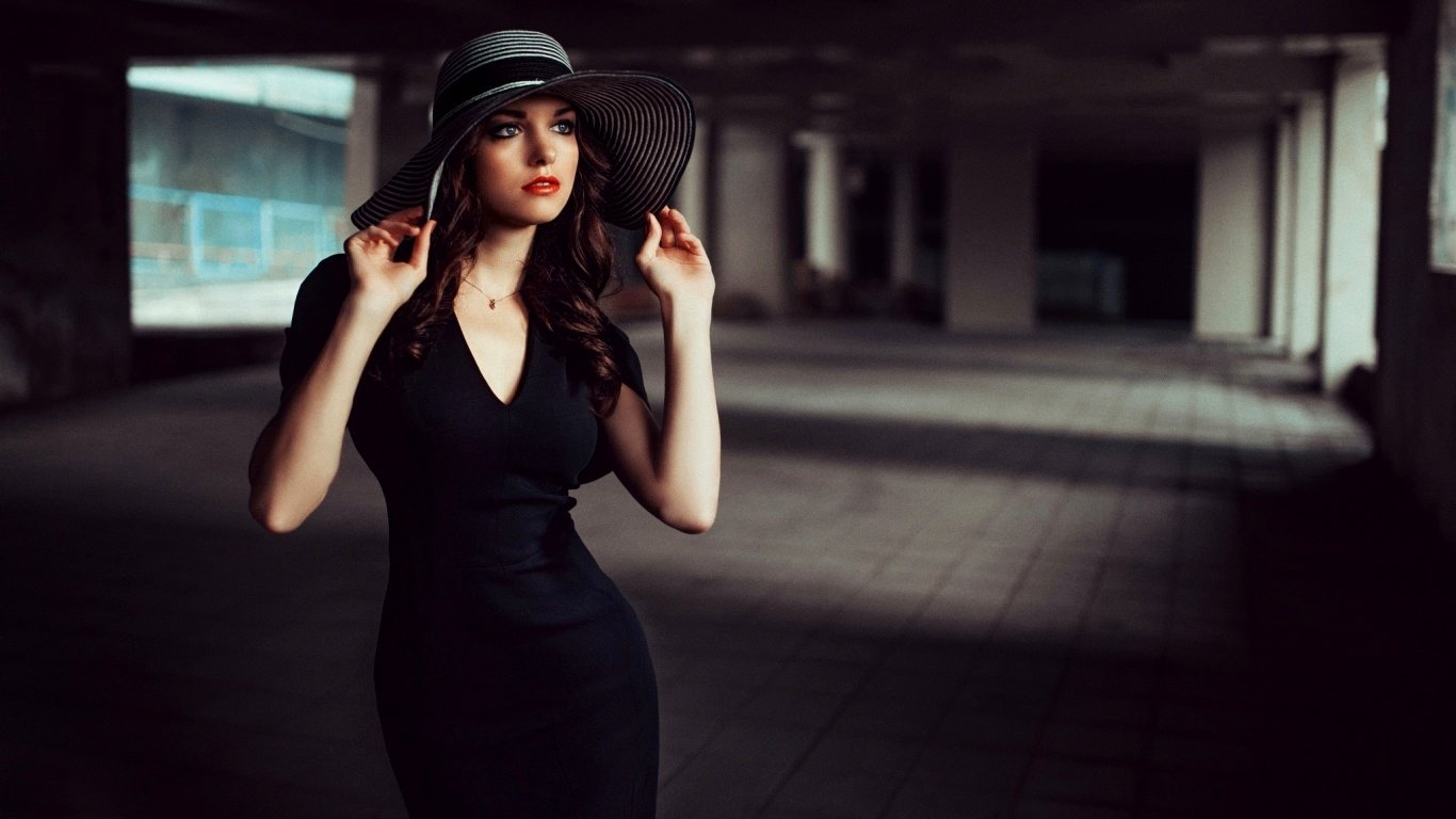 A confident woman model wearing a black dress and wide-brimmed hat poses in a dimly lit, spacious urban setting.