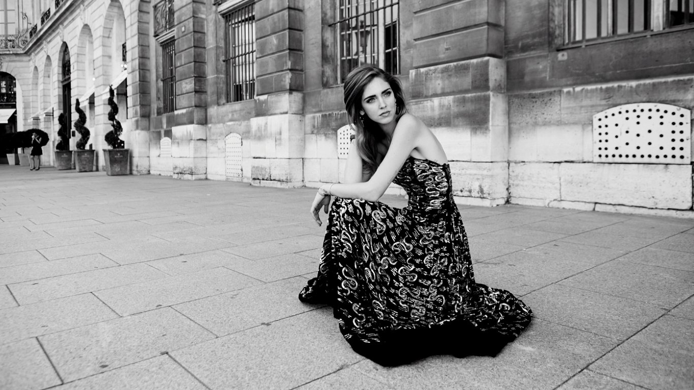 A woman named Chiara Ferragni sits gracefully on the ground, wearing an elegant black and floral dress, against the backdrop of a stylish urban setting in black and white.