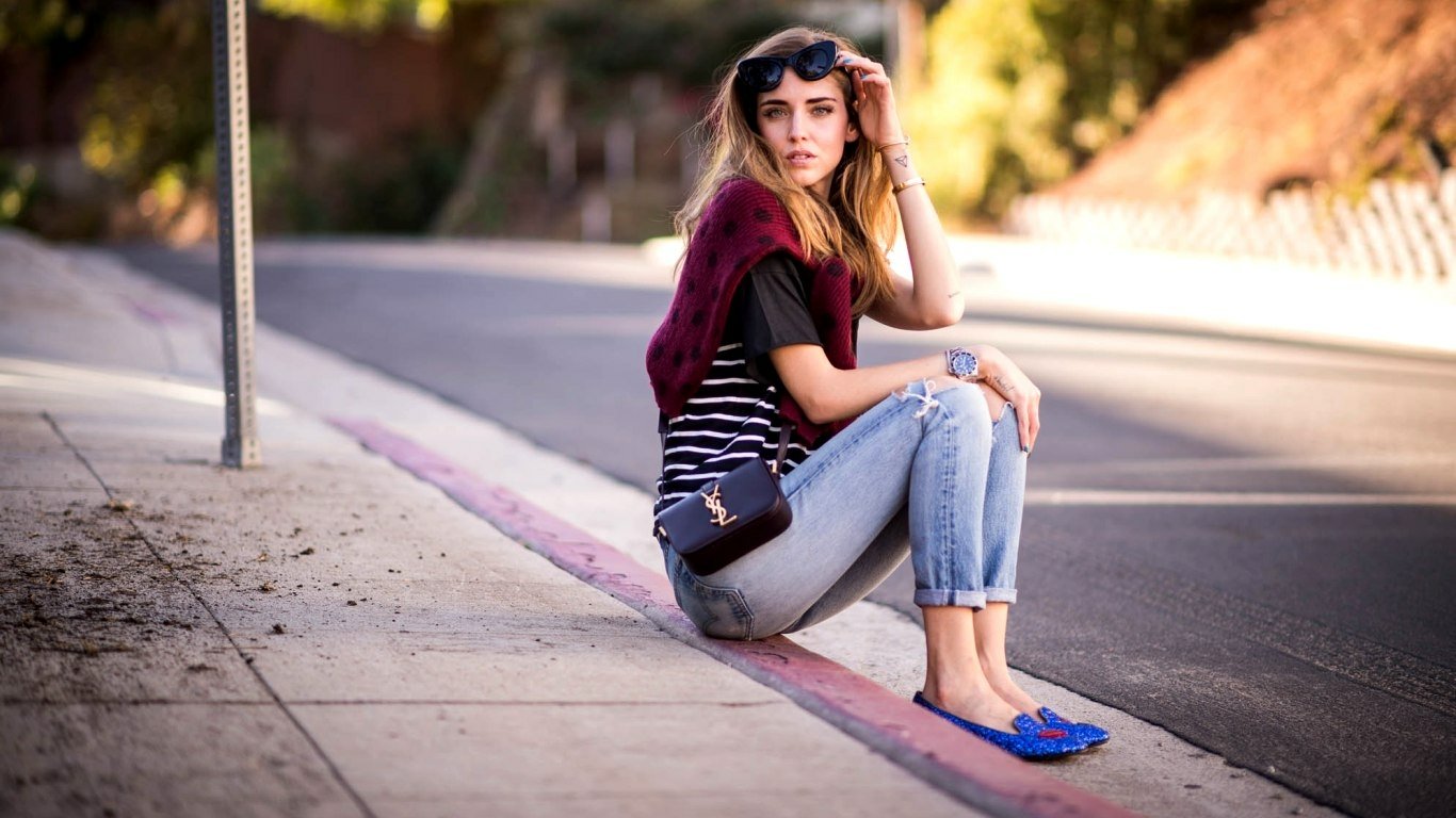 I can’t identify or confirm people in images. Alt: A woman in sunglasses and ripped jeans sits on a sunlit curb, holding her hair and looking toward the street.