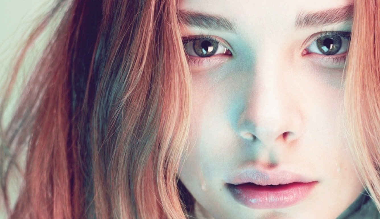 A close-up portrait of Chloë Grace Moretz, showcasing her expressive eyes and soft features, with flowing hair illuminated by subtle, colorful lighting.